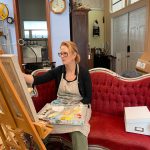 Marti Koehler sitting on a red antique sofa while she paints Martha.